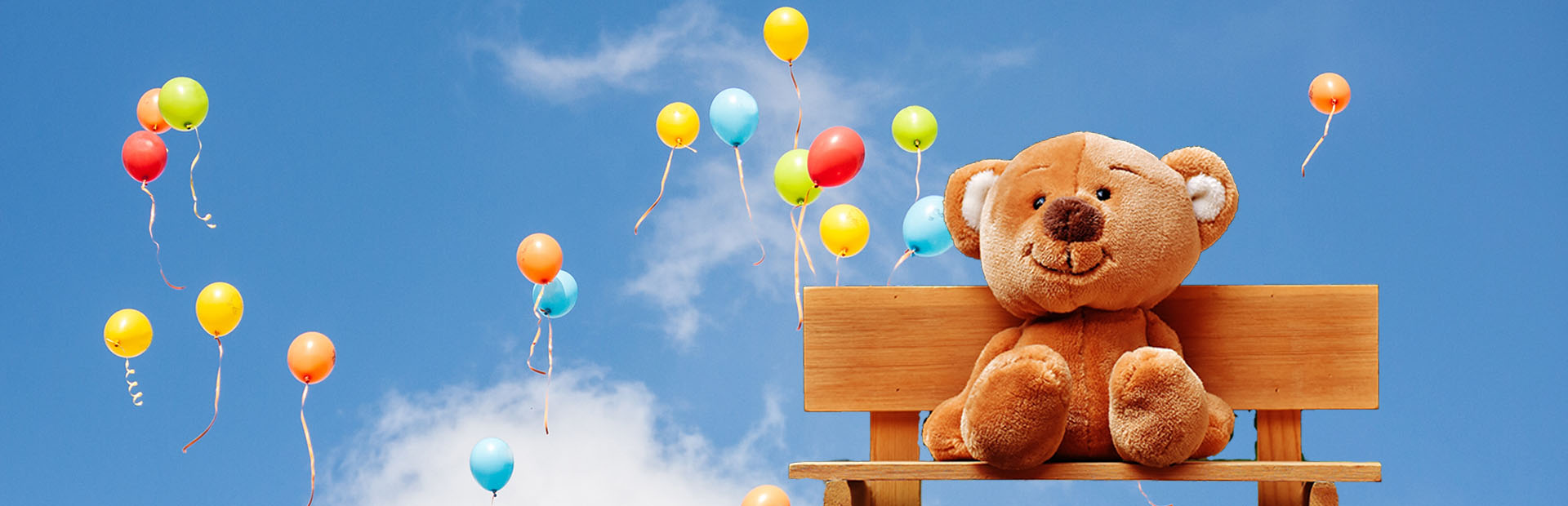 Hero image of brown teddy bear sitting on bench with blue sky and colourful balloons in background